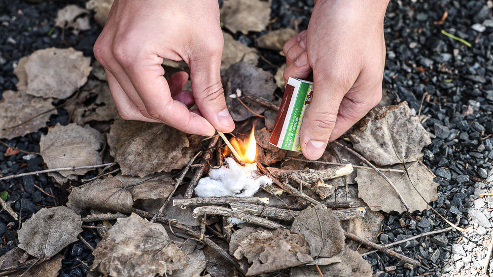 Hand's using a lighter to ignite the Coghlan's tinder over wood and twigs